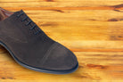 A Charcoal suede Oxford cap-toe shoe with a leather sole, displayed on a wooden surface.