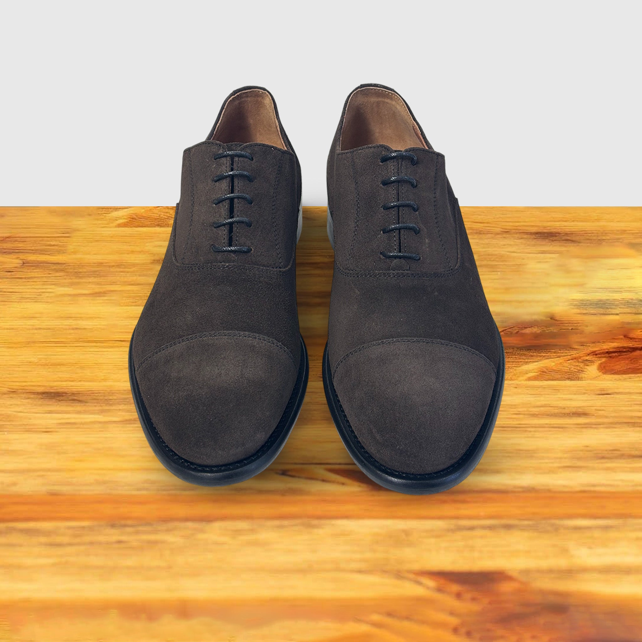 A Pair of  Charcoal suede Oxford cap-toe shoe with a leather sole, displayed on a wooden surface.