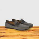 A Pair of  suede penny loafer with a strap, in a gray color, displayed on a wooden surface.