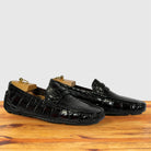 A pair of burgundy Genuine Crocodile leather driving loafers with gold-tone metal shoe trees inside, displayed on a wooden surface against a light gray background.