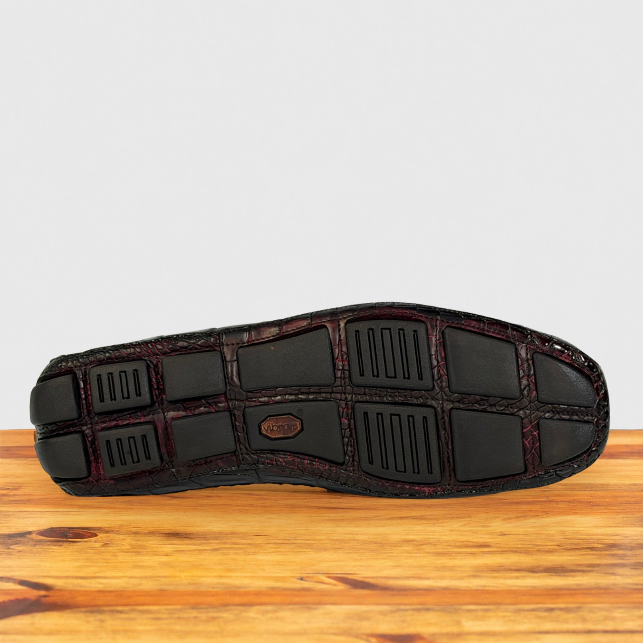The outsole of a shoe shown from the bottom, featuring black molded tread blocks and a textured dark burgundy base, resting on a wooden surface against a light gray background.