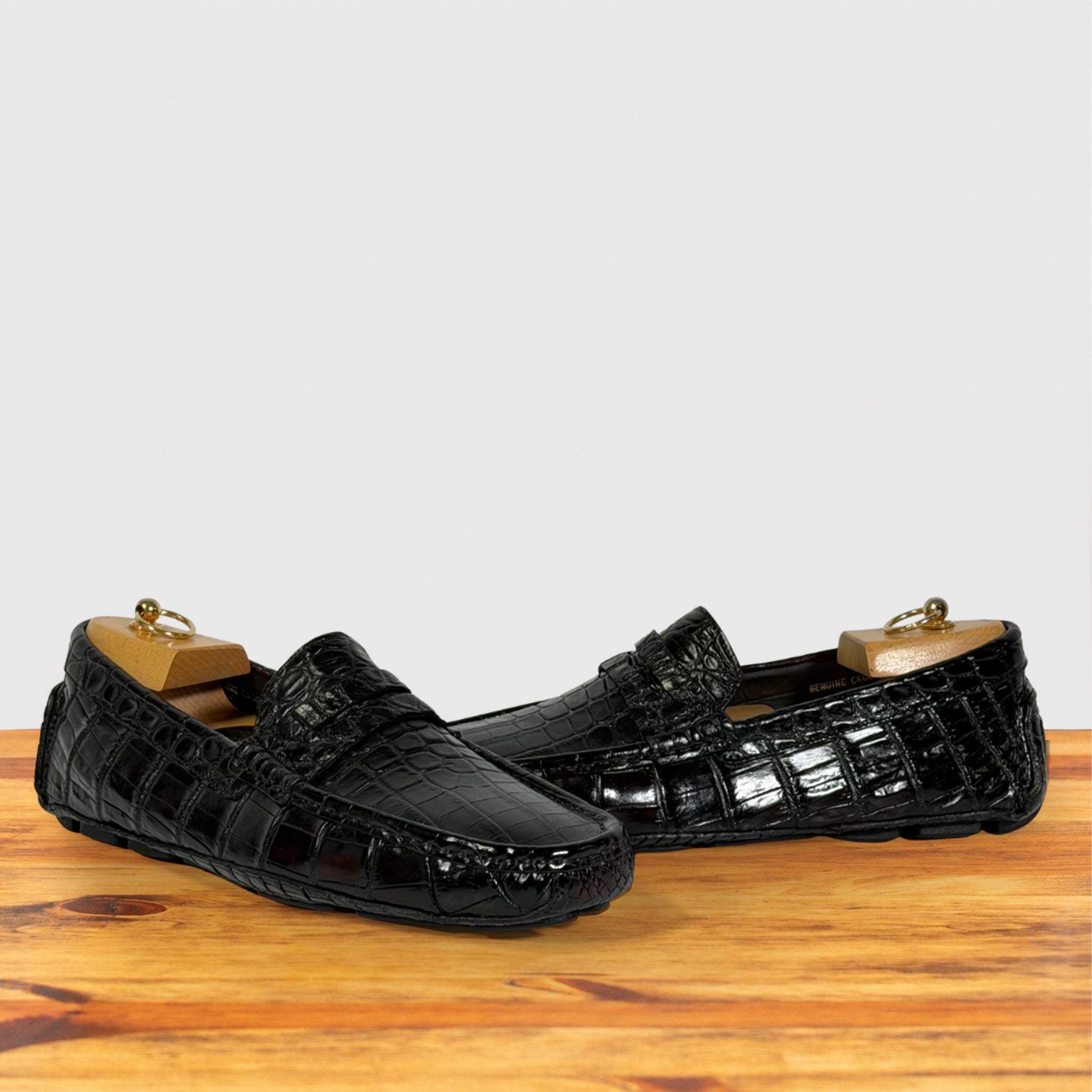 A pair of burgundy Genuine Crocodile leather penny loafers, displayed on wooden shoe trees on a wooden surface against a light gray background.