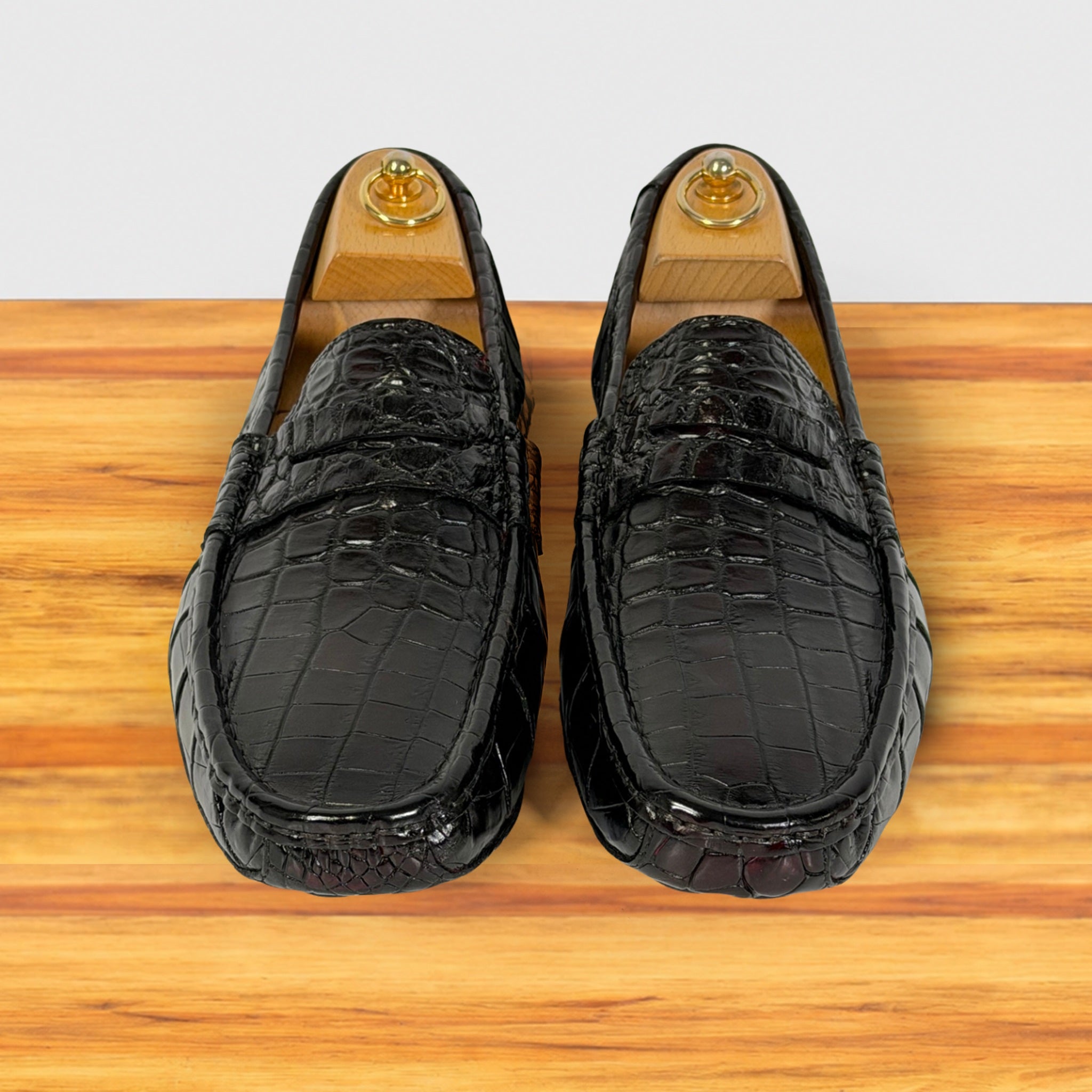 A pair of burgundy Genuine Crocodile leather loafers displayed on wooden shoe trees over a light wood surface against a white background.