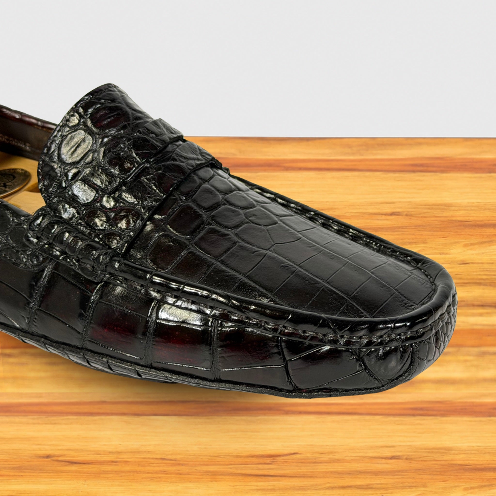 A burgundy Genuine Crocodile leather loafer shown in a close-up side view, resting on a light wood surface against a white background.