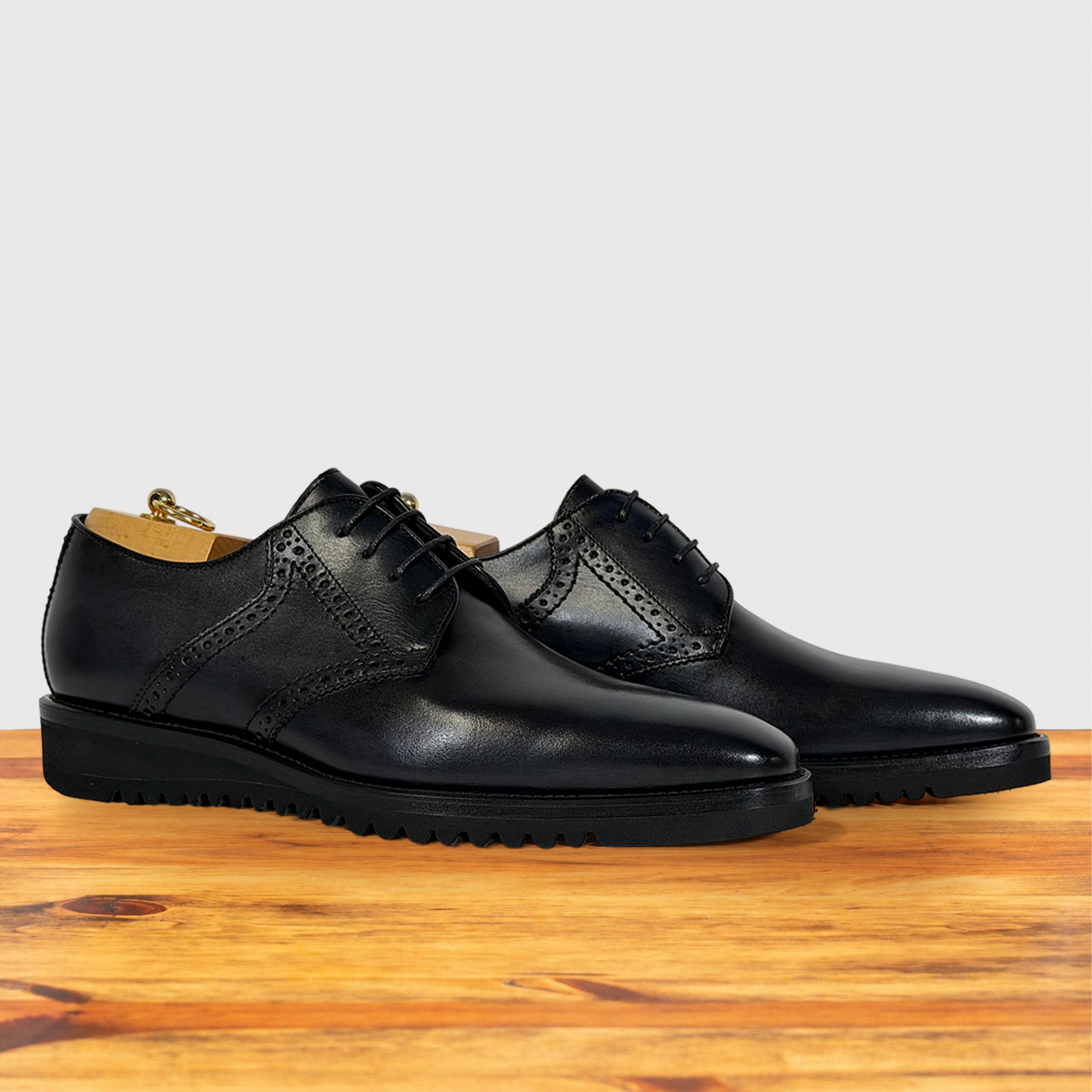 A pair of men's graphite leather oxford shoes with brogue detailing with laces, displayed on a wooden surface against a light gray background.