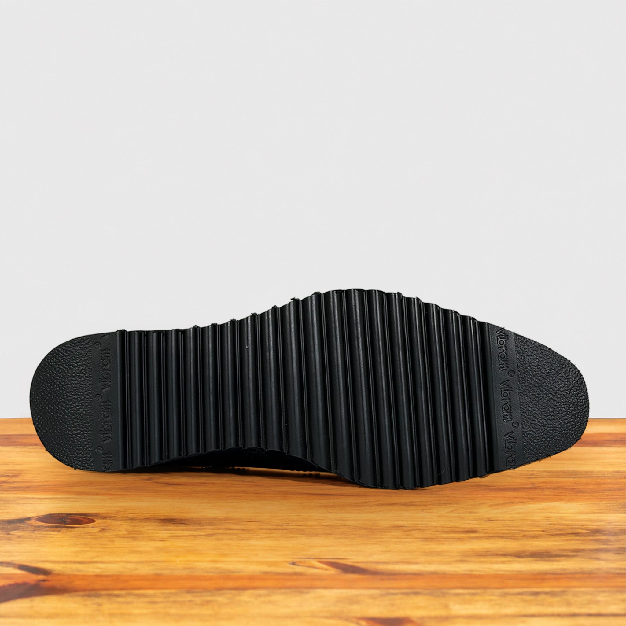 The black textured rubber outsole of a shoe, showing deep channeling and tread details, resting on a wooden surface against a light gray background.