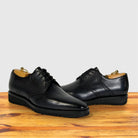 A pair of graphite leather lace-up dress shoes with brogue detailing, displayed on a wooden surface against a light gray background.