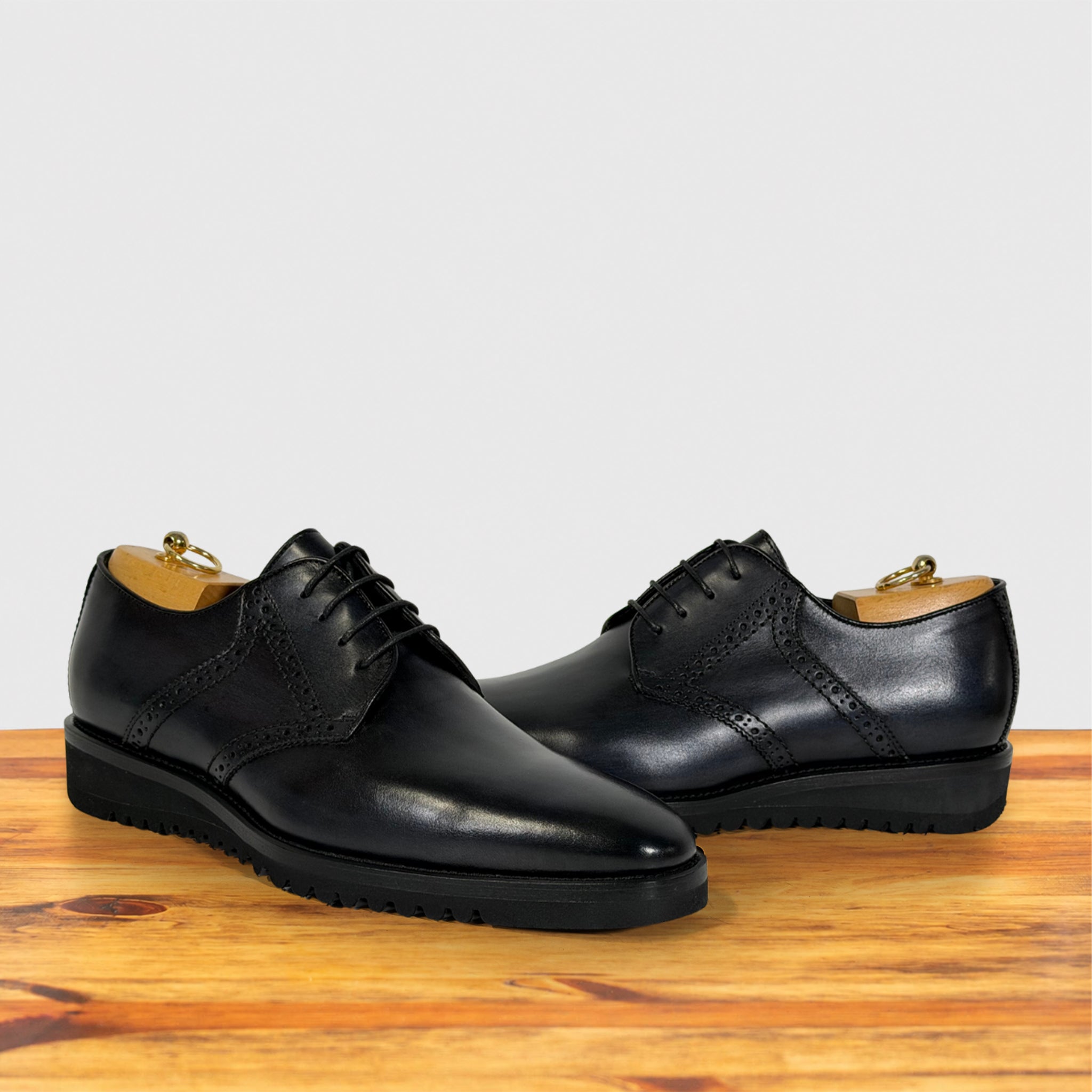 A pair of graphite leather lace-up dress shoes with brogue detailing, displayed on a wooden surface against a light gray background.