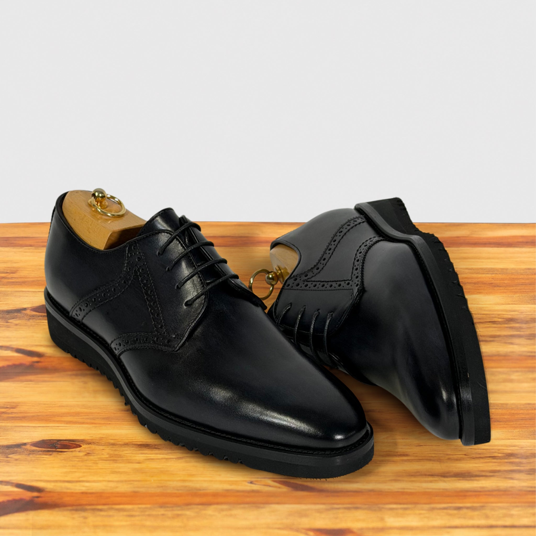 A pair of graphite leather oxford dress shoes with brogue detailing, displayed on a wooden surface against a light gray background.