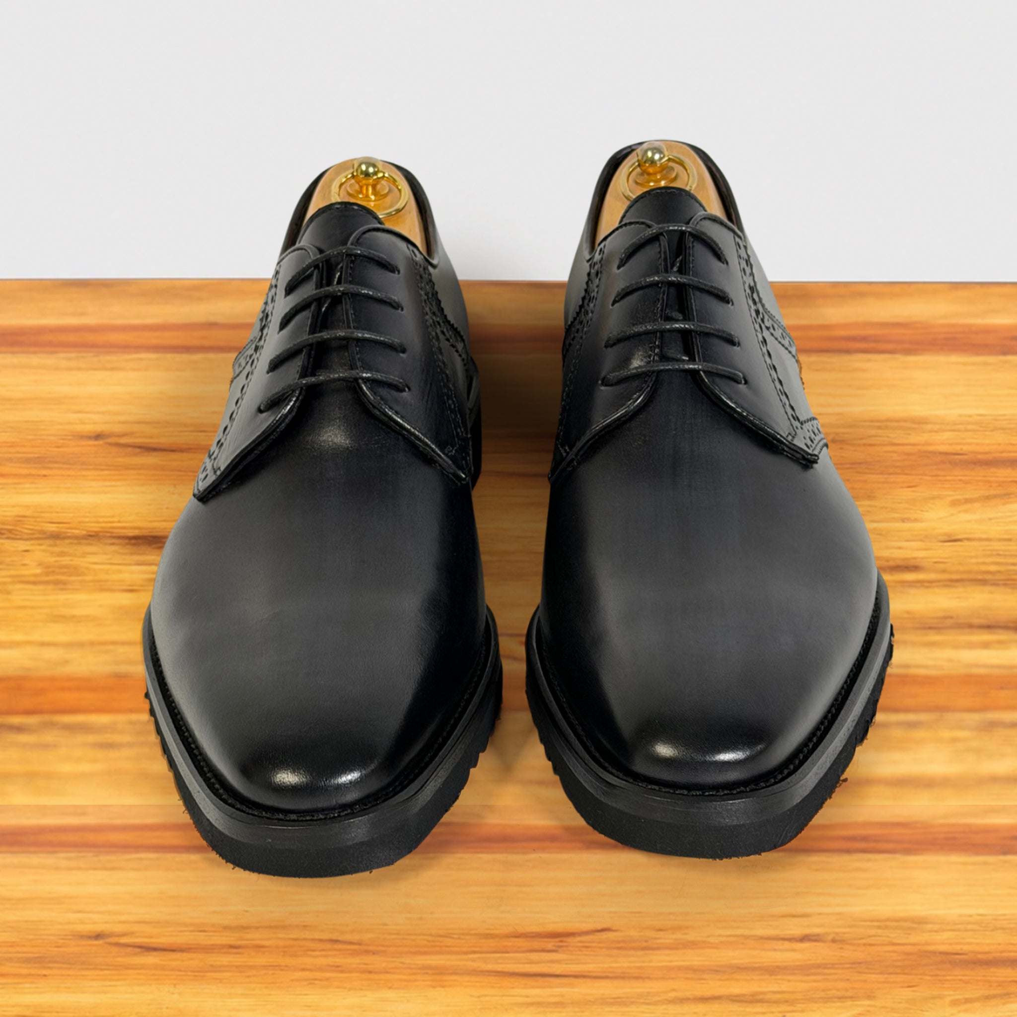 A pair of men's graphite leather oxford shoes with brogue detailing, displayed from a front-facing perspective on a wooden surface against a white background.