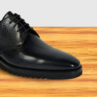 A close-up of a graphite leather lace-up dress shoe with brogue detailing, resting on a light brown wooden surface against a white background.