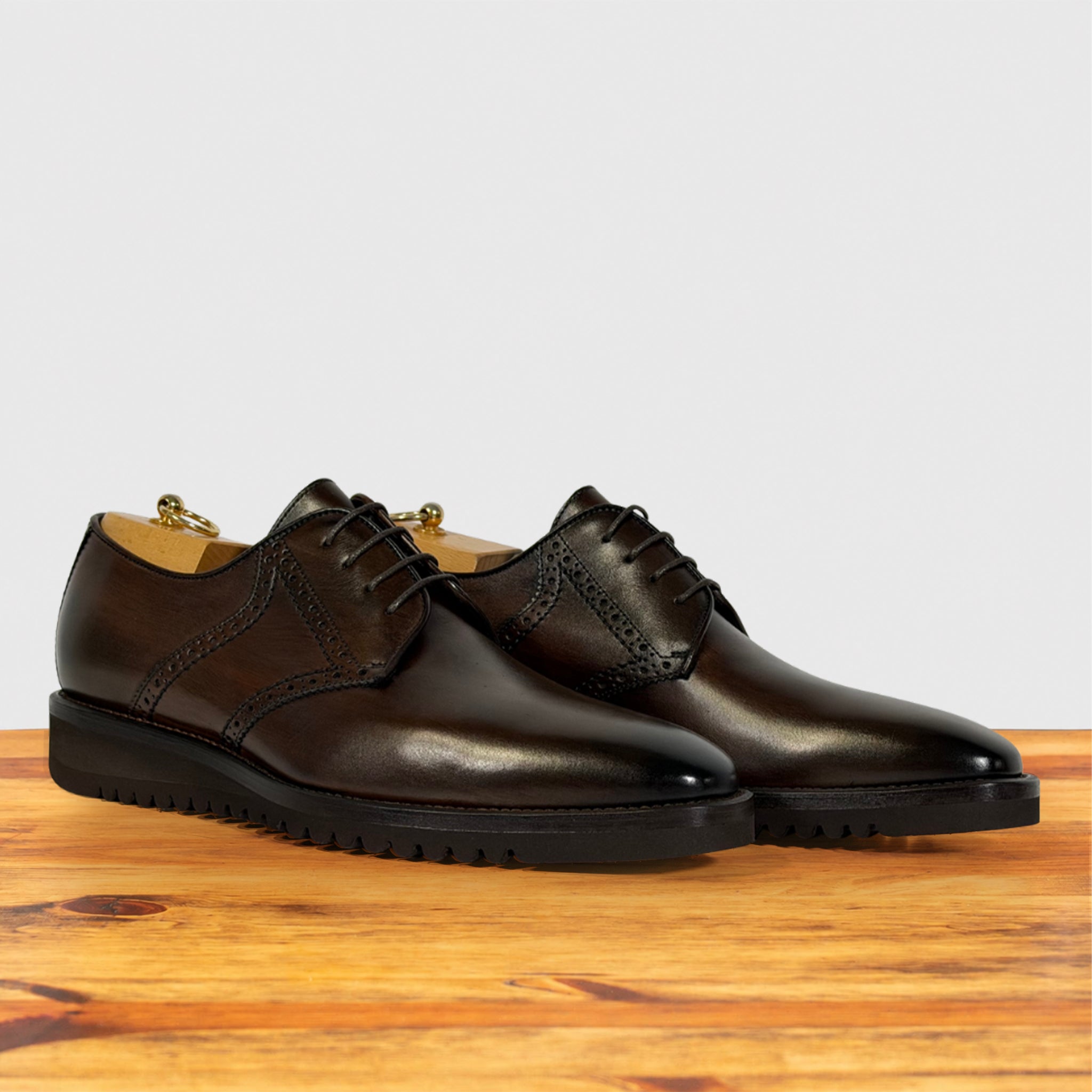 A pair of  moor leather oxford shoes with brogue detailing and black soles, displayed on a wooden surface against a light gray background.