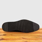 The black textured rubber outsole of a shoe, showing ribbed tread details, resting on a wooden surface against a light gray background.