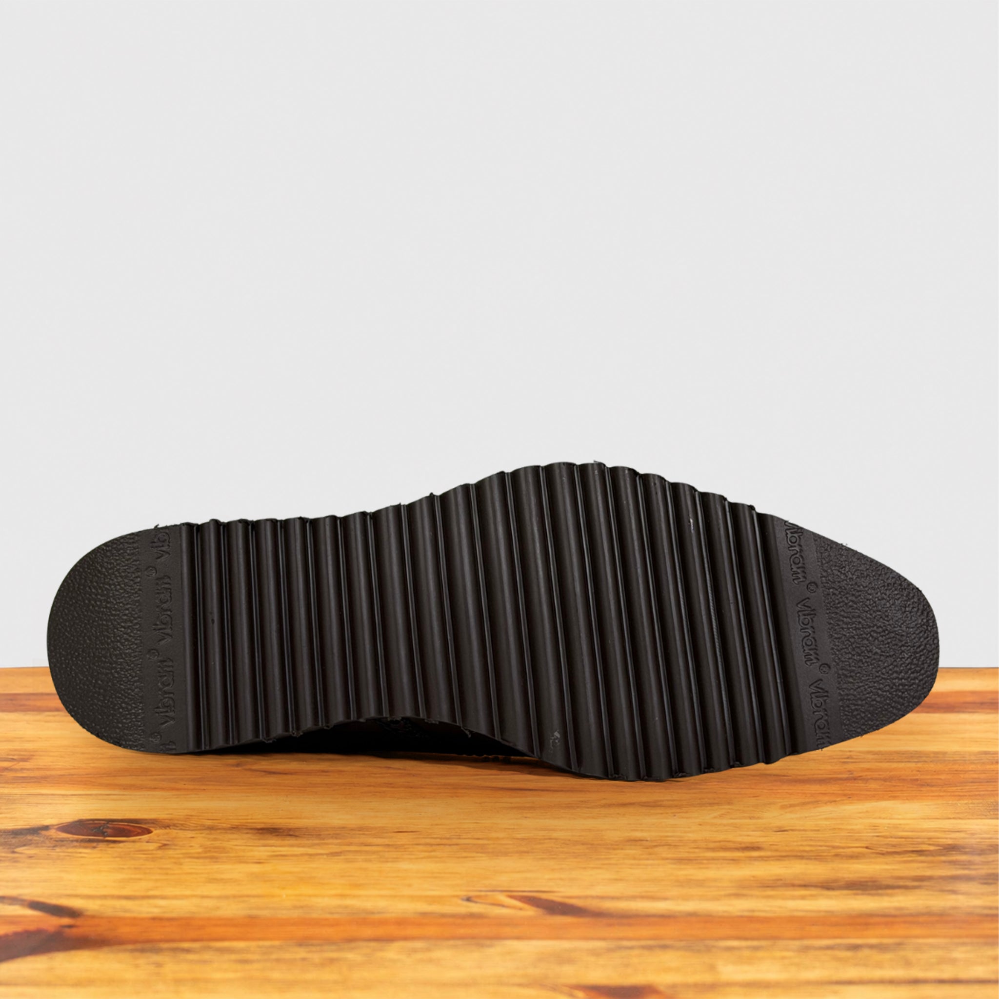 The black textured rubber outsole of a shoe, showing ribbed tread details, resting on a wooden surface against a light gray background.
