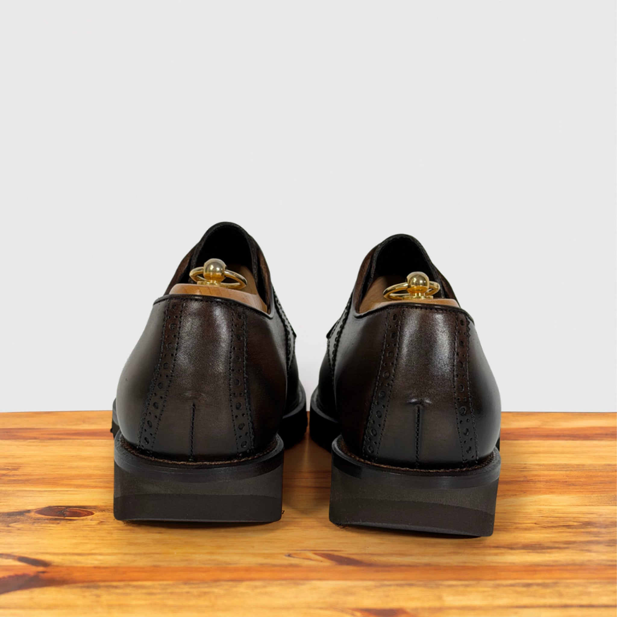 A pair of moor leather dress shoes shown from the back, resting on a wooden surface against a light gray background.
