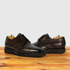A pair of moor leather lace-up dress shoes with brogue detailing and black soles, displayed on a wooden surface against a light gray background.
