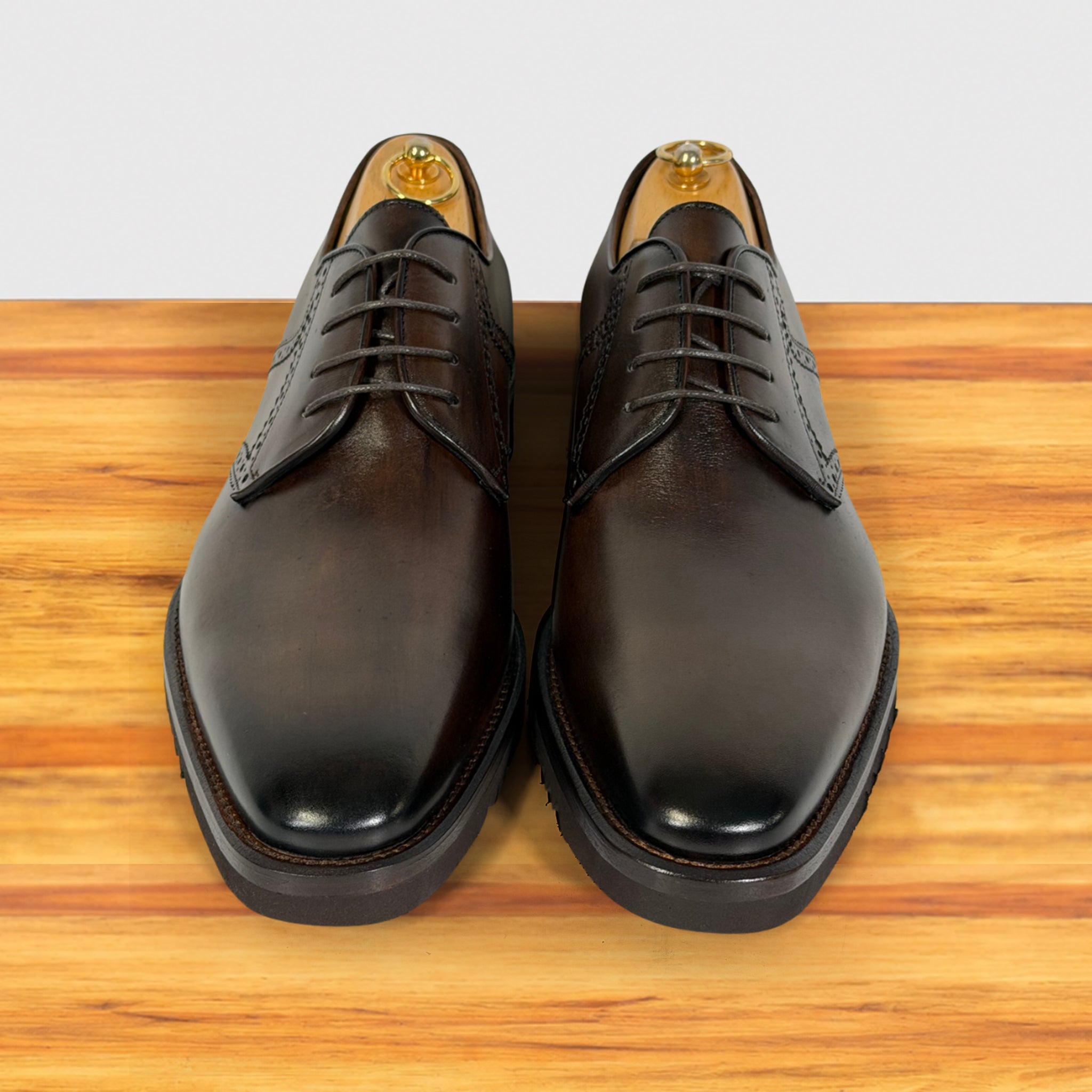 A pair of moor leather oxford dress shoes with brogue detailing, displayed on a wooden surface against a light gray background.