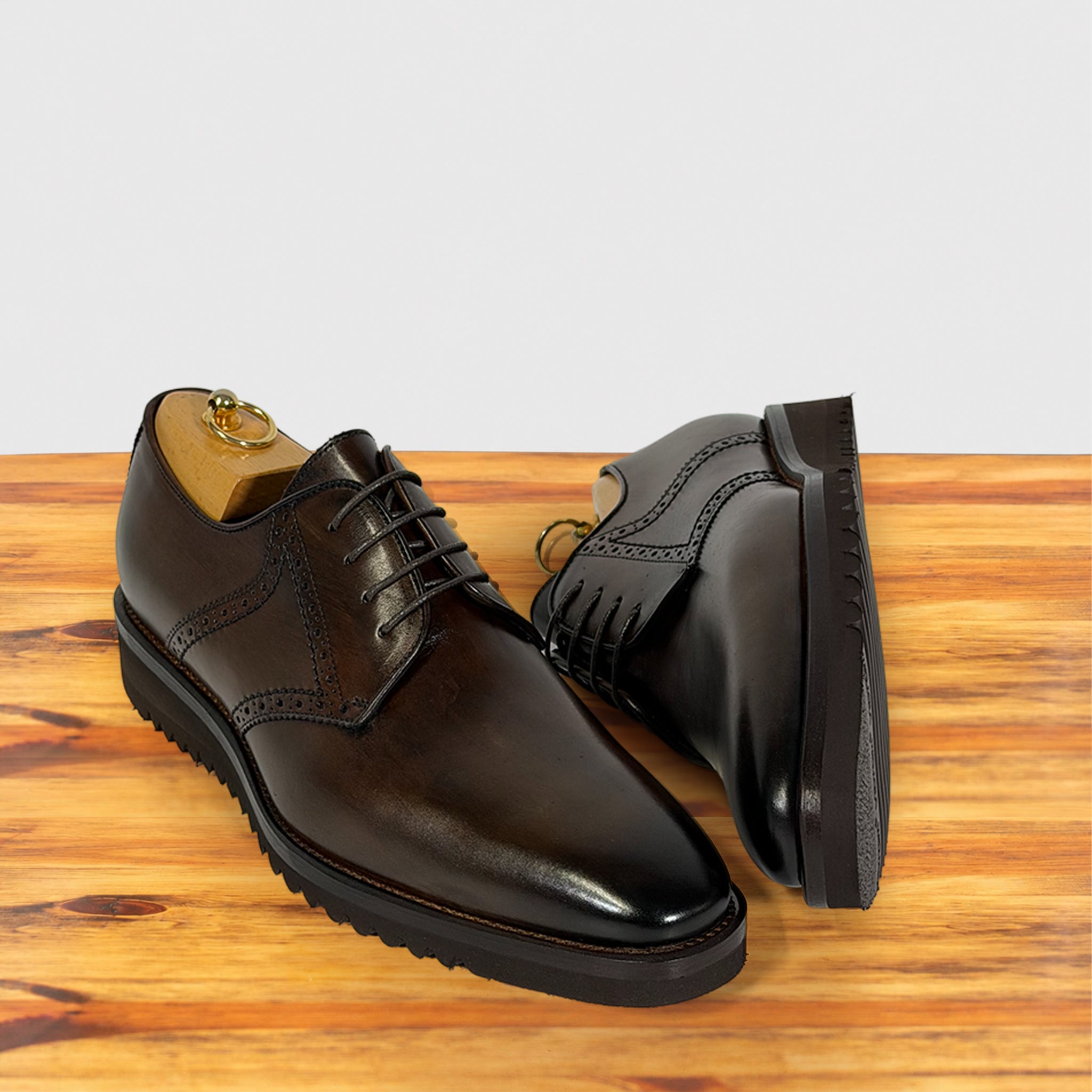 A pair of moor leather lace-up dress shoes with brogue detailing, displayed on a wooden surface against a light gray background.