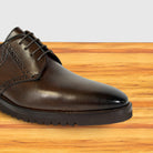 A close-up of a moor leather lace-up dress shoe with decorative brogue detailing and a rugged black sole, shown on a wooden surface against a light gray background.