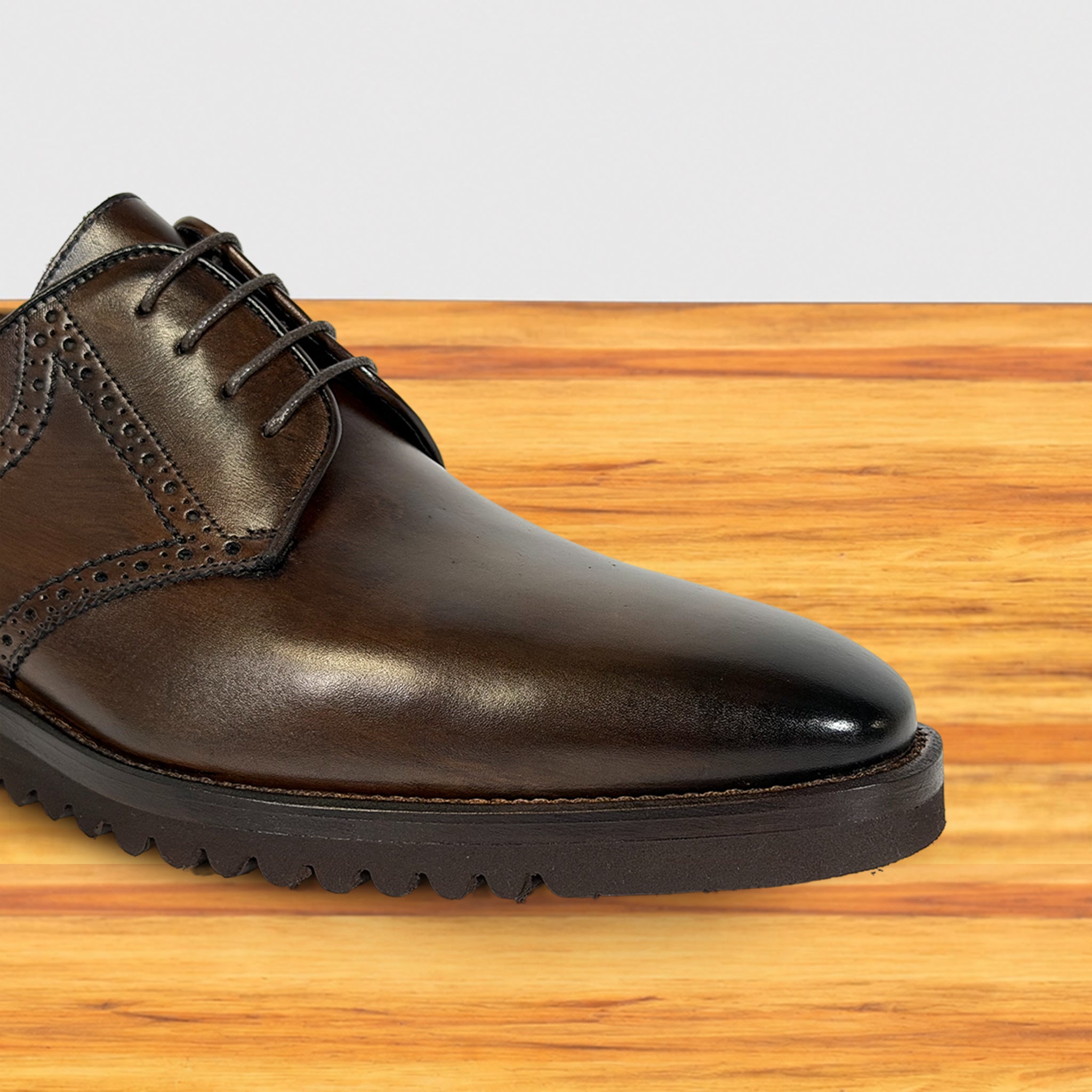 A close-up of a moor leather lace-up dress shoe with decorative brogue detailing and a rugged black sole, shown on a wooden surface against a light gray background.