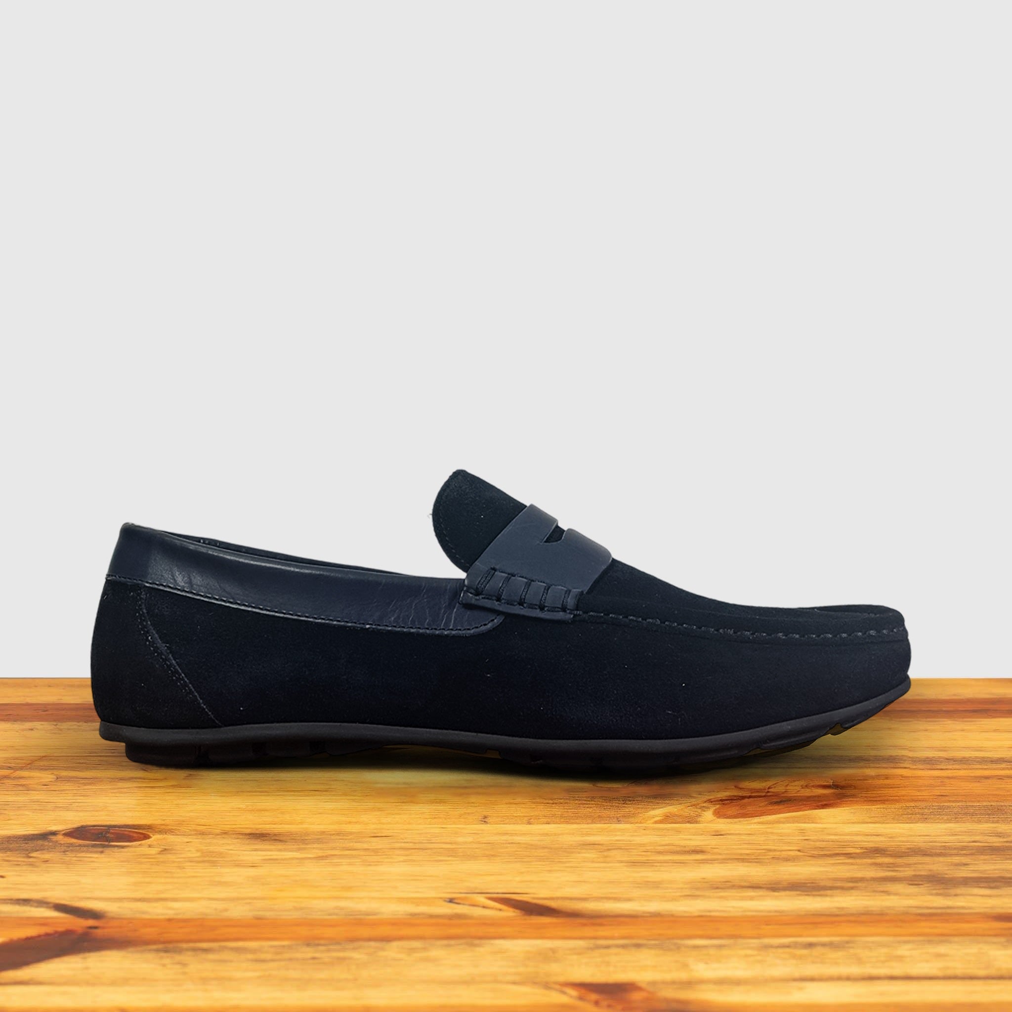 A navy blue suede penny loafer with a  strap, displayed on a wooden surface.