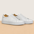 A pair of white leather low-top sneakers with perforated side panels, white laces, displayed on a light wood surface against a warm beige background.
