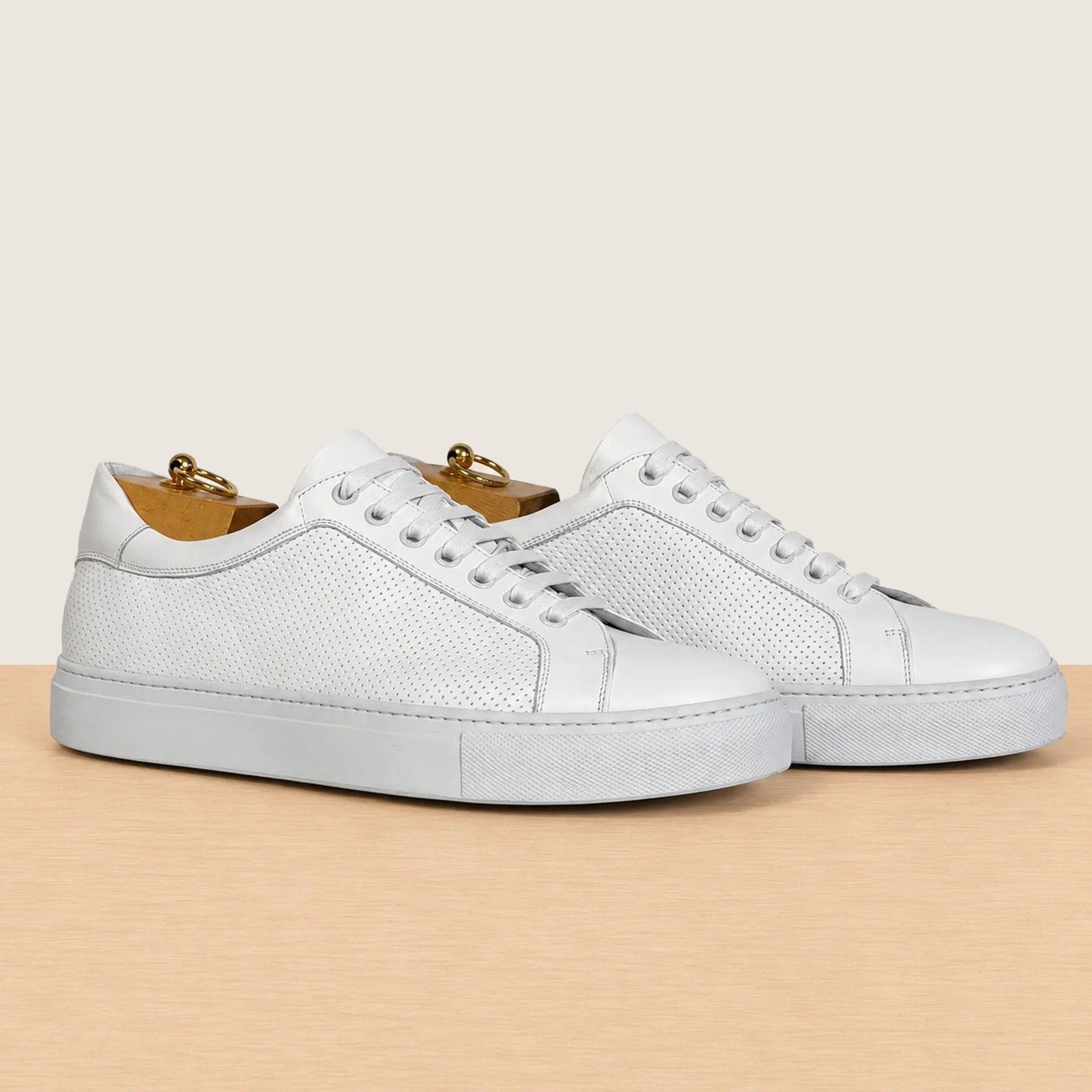 A pair of white leather low-top sneakers with perforated side panels, white laces, displayed on a light wood surface against a warm beige background.