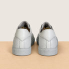 A pair of white leather low-top sneakers shown from the back, positioned side-by-side on a light wood surface against a solid beige background.