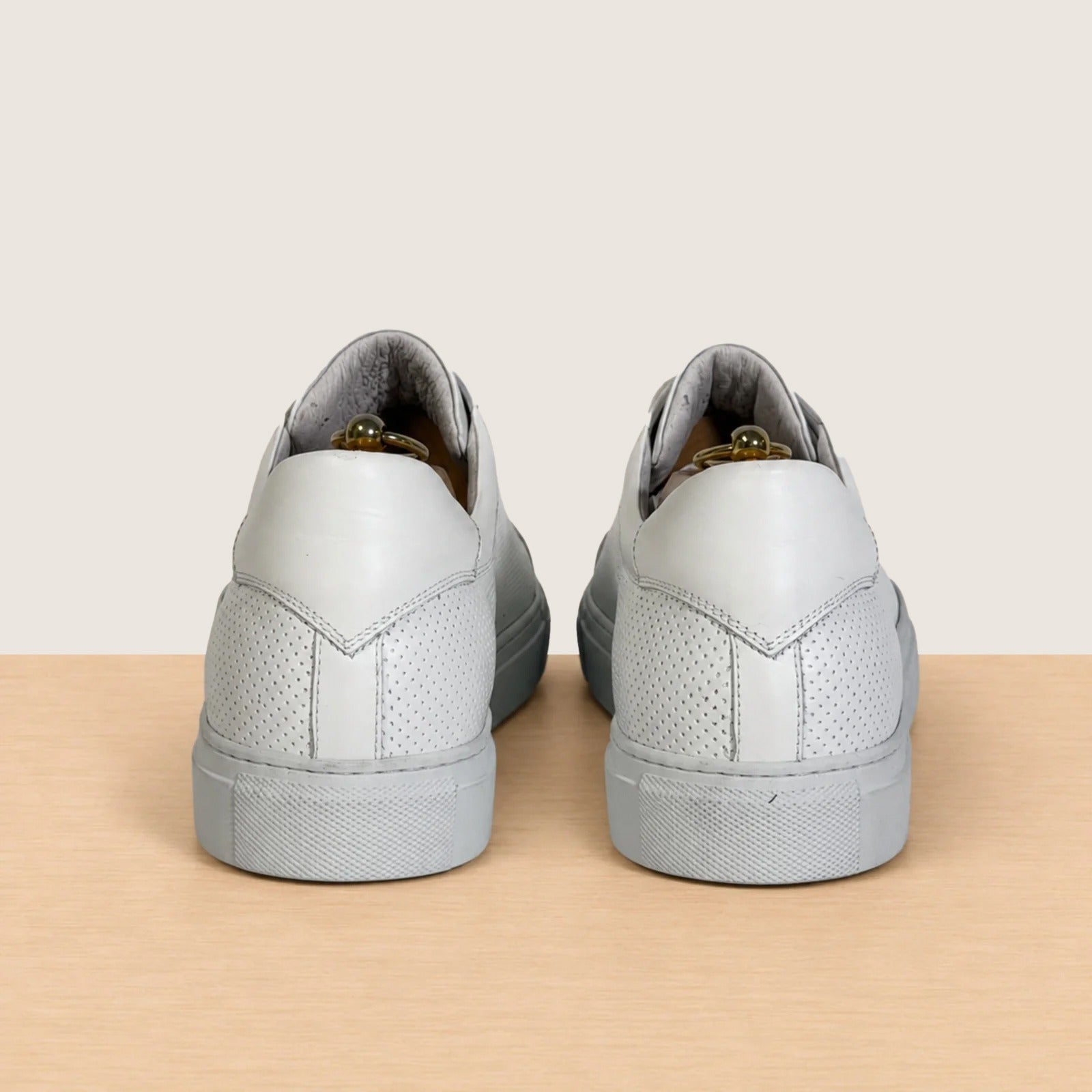 A pair of white leather low-top sneakers shown from the back, positioned side-by-side on a light wood surface against a solid beige background.