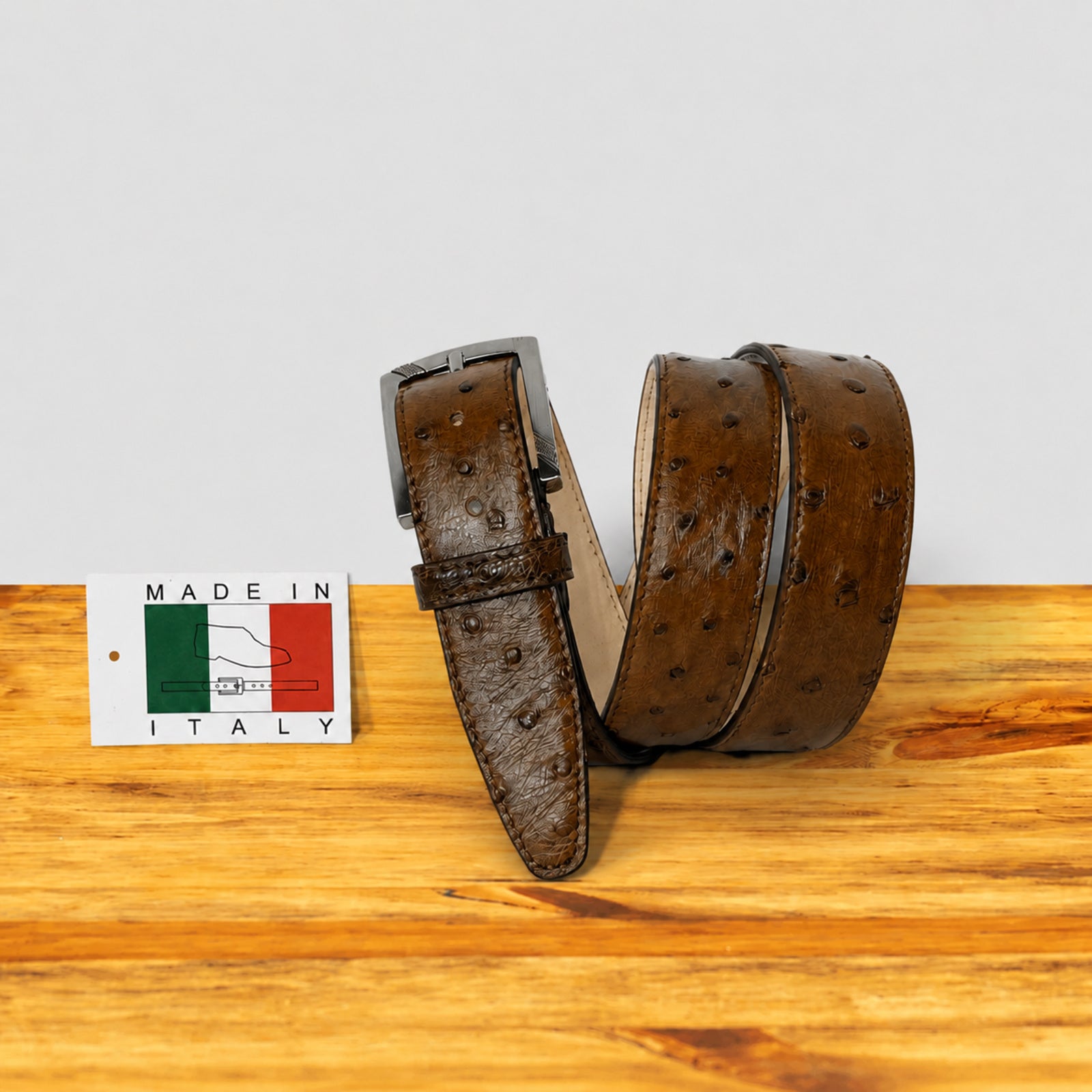 A Cerris ostrich leather belt with a metal buckle, coiled on a wooden surface next to a small 'Made in Italy' tag.