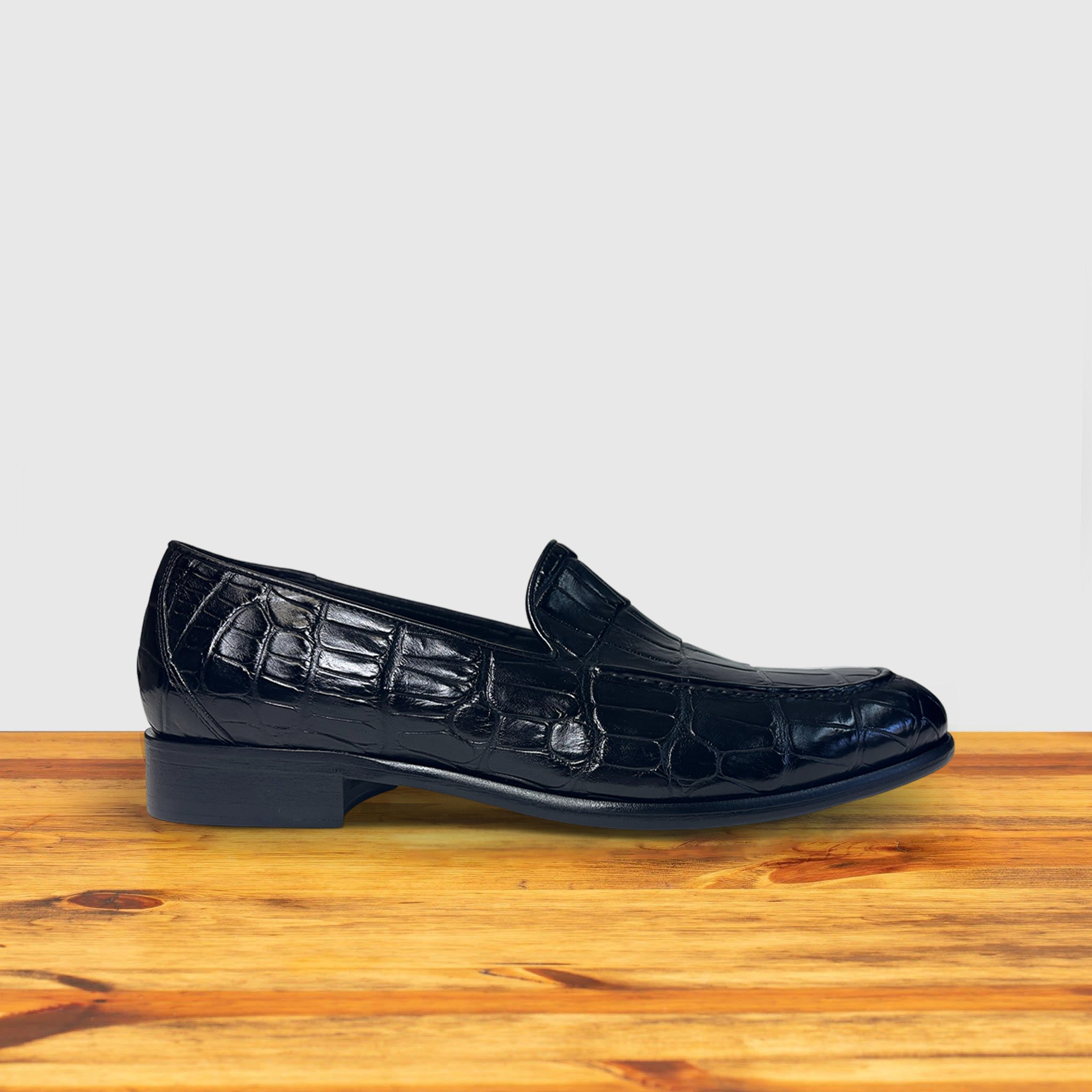 Black crocodile loafer shoe on a wooden surface with a light gray background