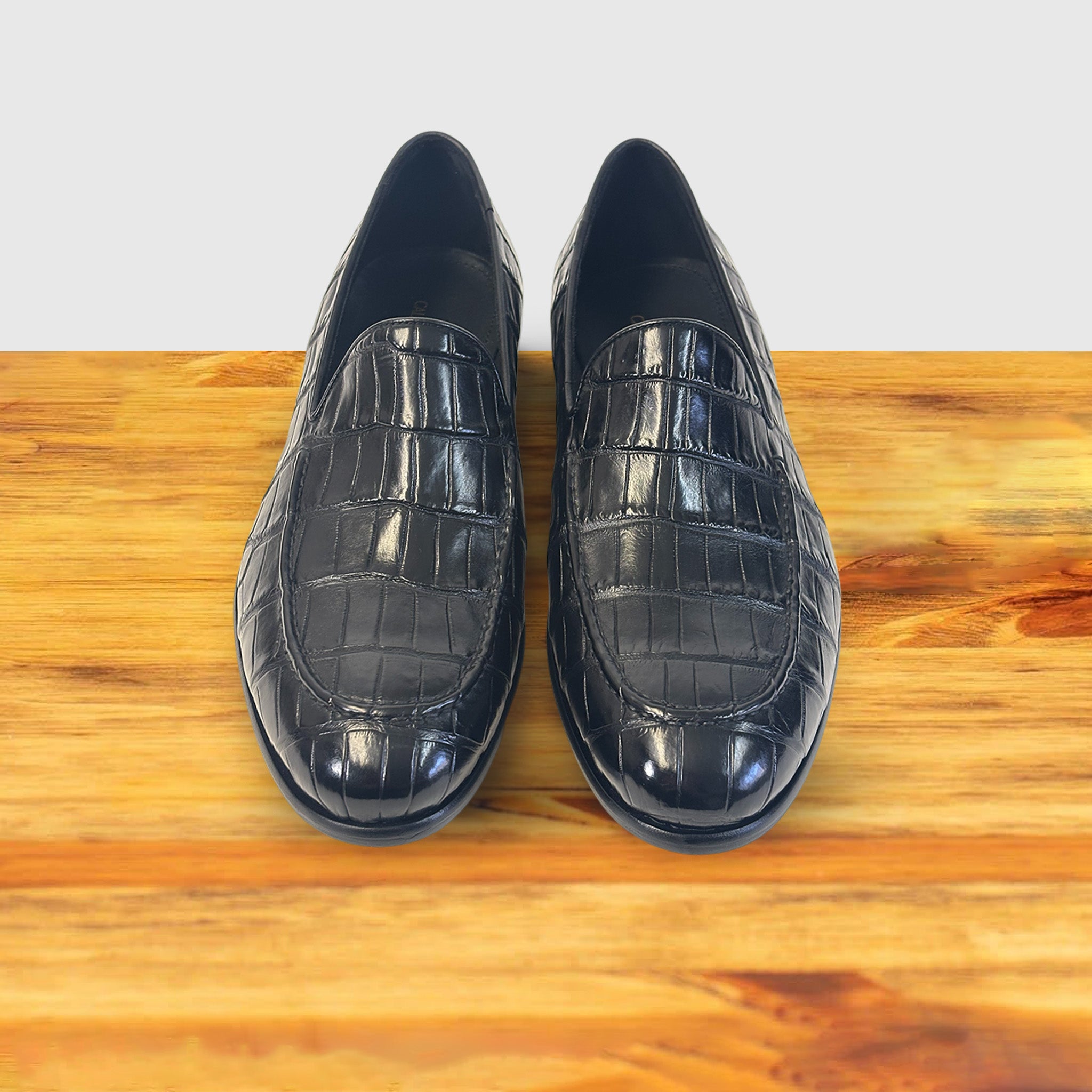 A pair of Black crocodile loafer shoe on a wooden surface with a light gray background