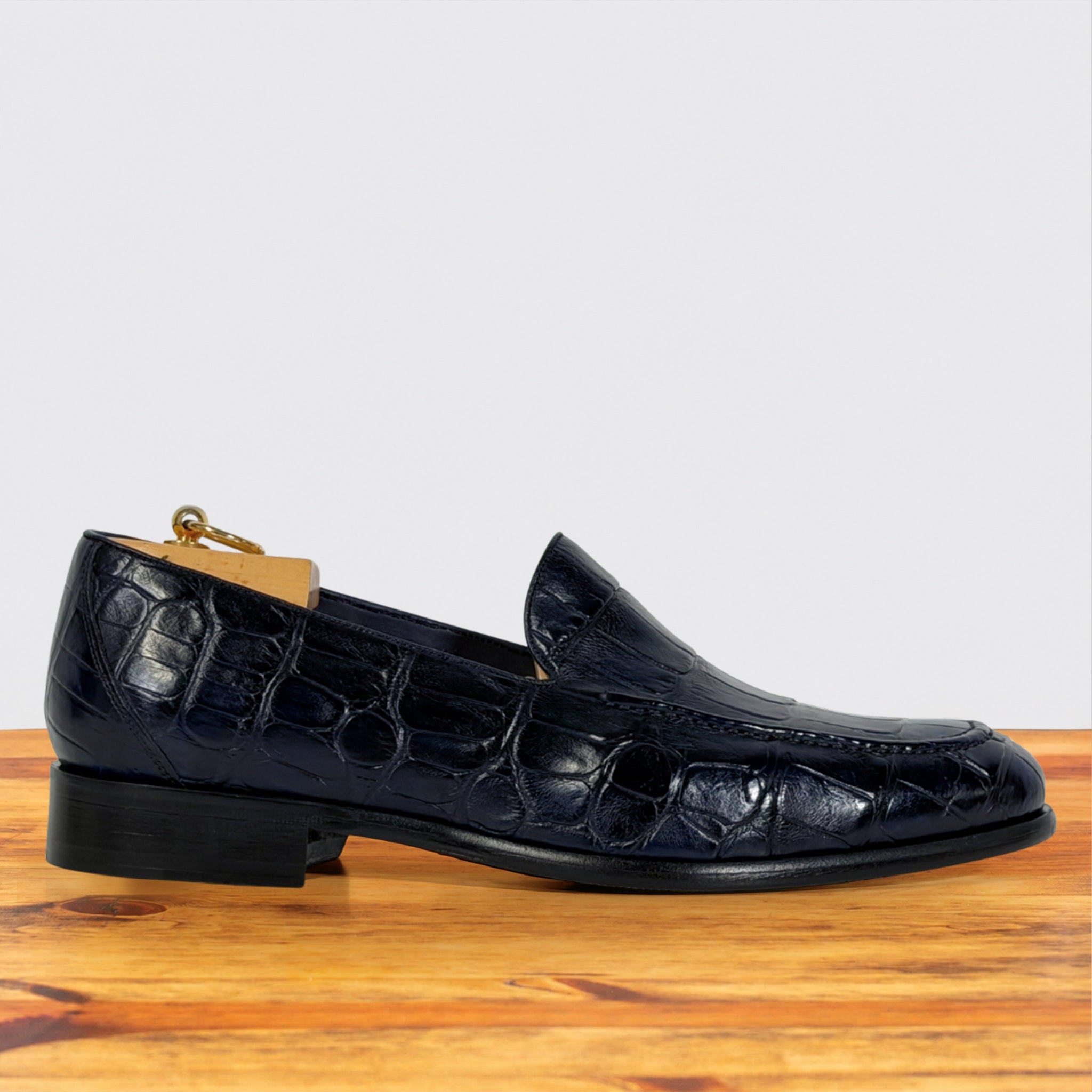 A single dark navy blue crocodile-embossed leather loafer shown in profile on a wooden surface against a light gray background.