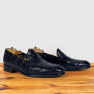 A pair of dark navy blue crocodile-embossed leather loafers resting on a wooden surface against a light gray background.