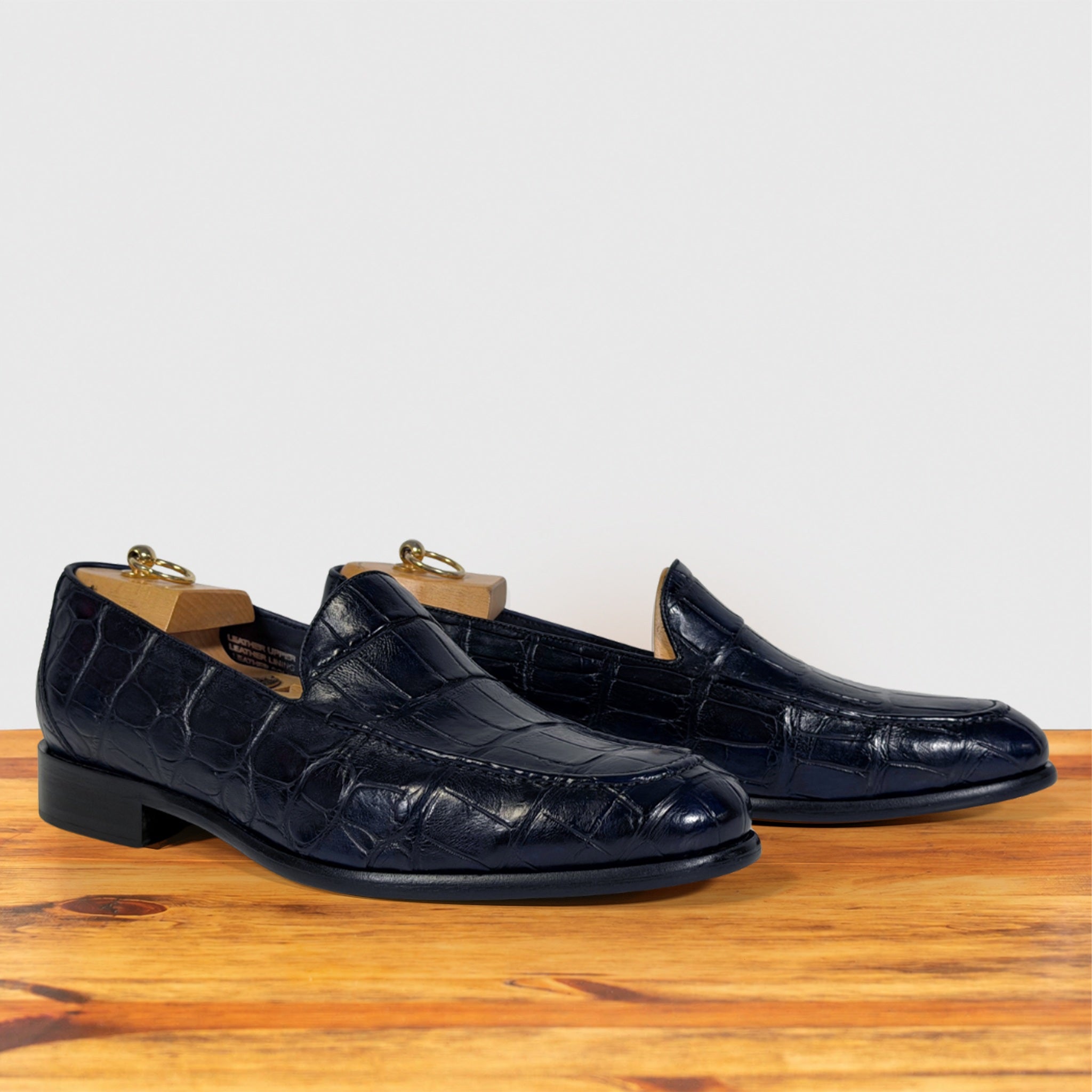 A pair of dark navy blue crocodile-embossed leather loafers resting on a wooden surface against a light gray background.