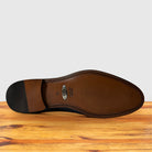 The outsole of a leather shoe is shown from a top-down view, resting on a wooden surface against a light gray background. The sole features a dark brown heel, a silver oval metal brand plate, and a stitched edge.