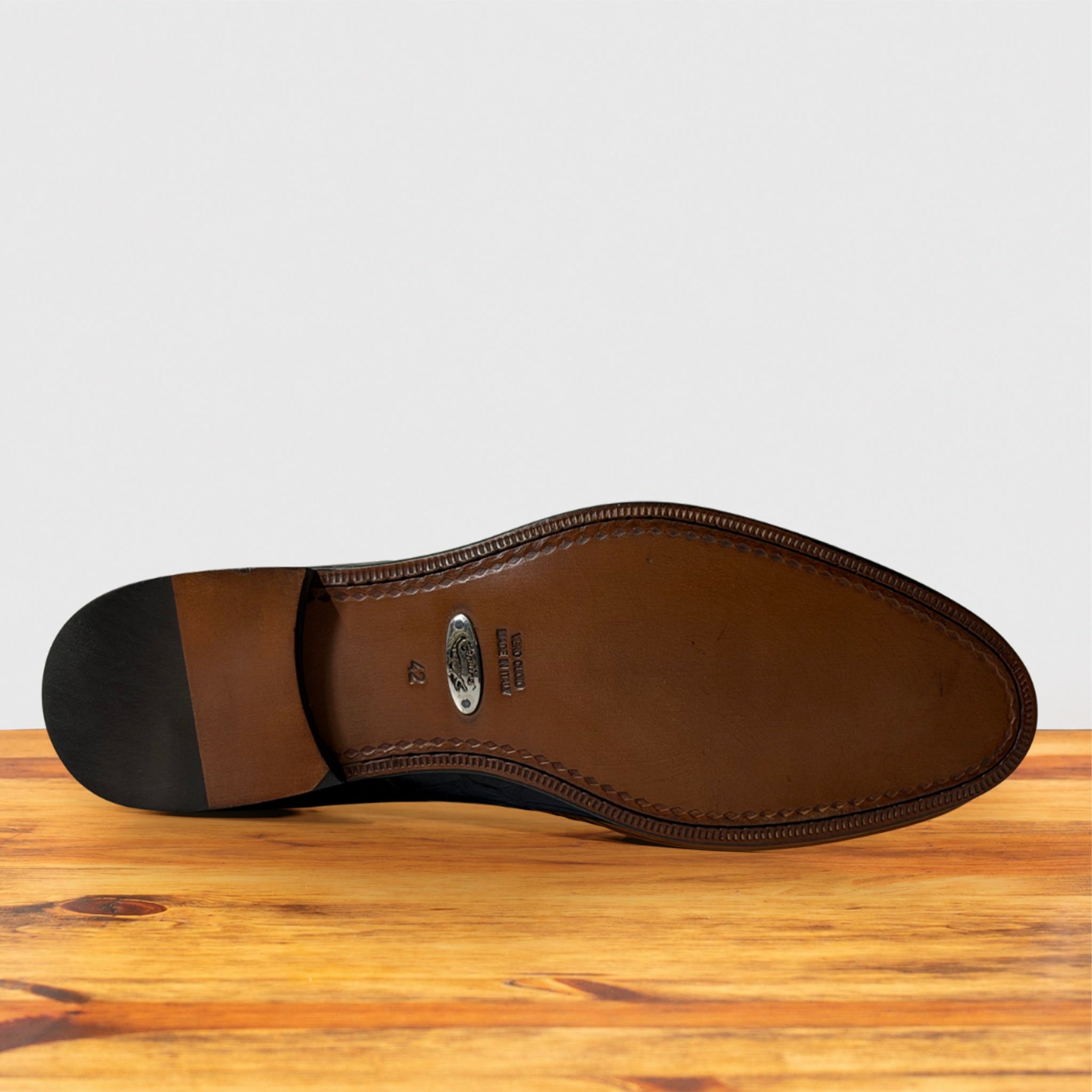 The outsole of a leather shoe is shown from a top-down view, resting on a wooden surface against a light gray background. The sole features a dark brown heel, a silver oval metal brand plate, and a stitched edge.