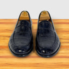 A pair of dark navy blue crocodile-embossed leather slip-on loafers with wooden shoe trees inserted, displayed on a wooden surface against a white background.