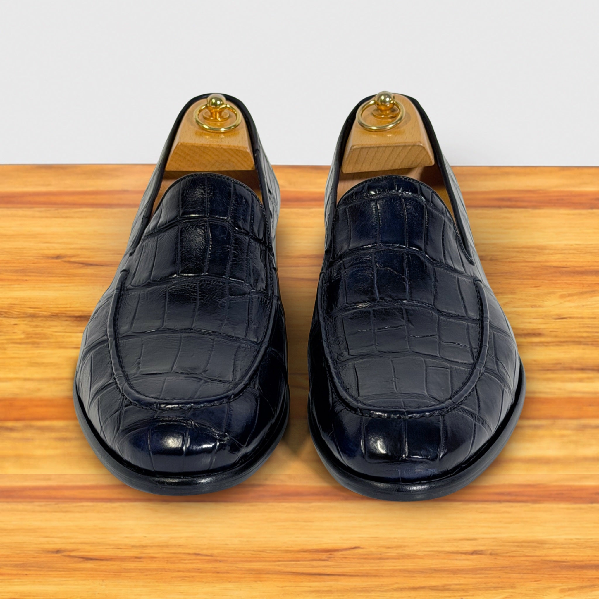 A pair of dark navy blue crocodile-embossed leather slip-on loafers with wooden shoe trees inserted, displayed on a wooden surface against a white background.