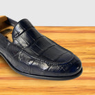 A single men's loafer in dark navy blue crocodile-embossed leather, shown resting on a light wood surface against a white background.