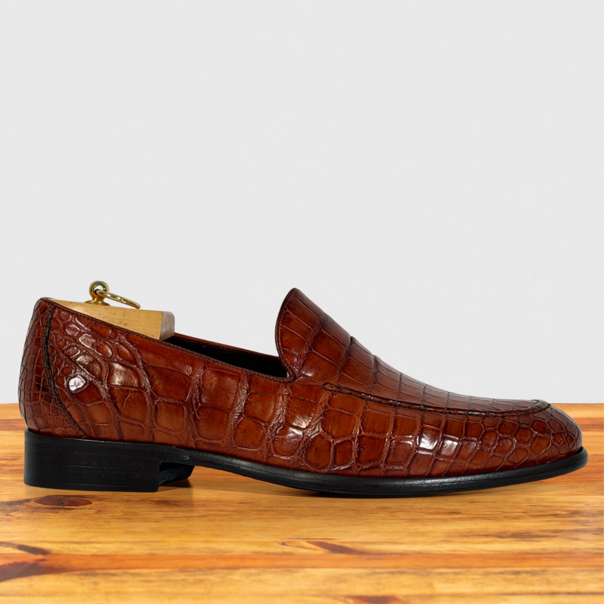 A single cognac crocodile-embossed leather loafer shown in profile on a wooden surface against a light gray background.