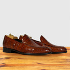 A pair of cognac crocodile-embossed leather loafers with black soles, displayed on a wooden surface against a light gray background.