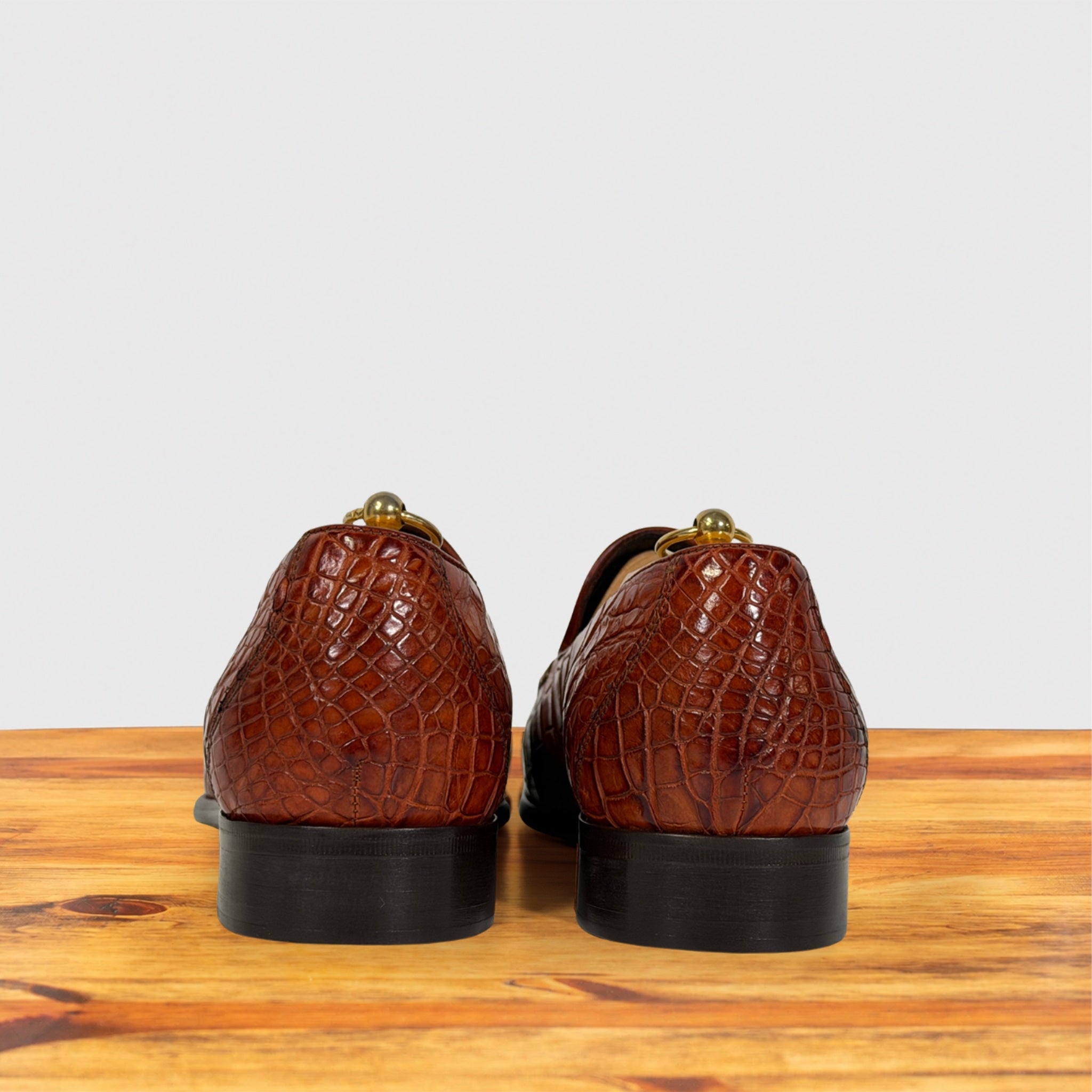 A pair of cognac crocodile-embossed leather loafers shown from the back, resting on a wooden surface against a light gray background.