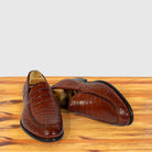 A pair of cognac crocodile-embossed leather loafers displayed on a wooden surface with a light gray background.