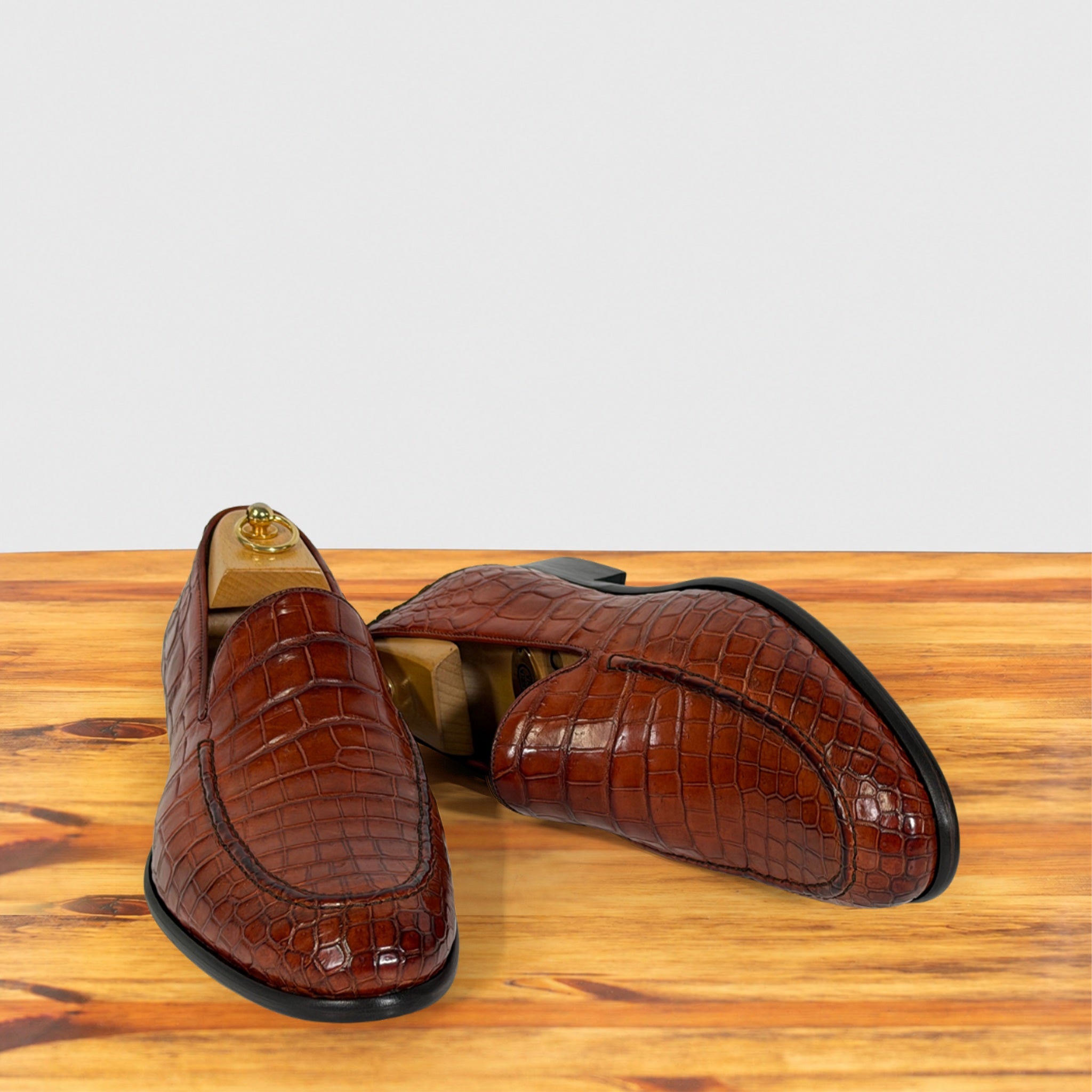A pair of cognac crocodile-embossed leather loafers displayed on a wooden surface with a light gray background.