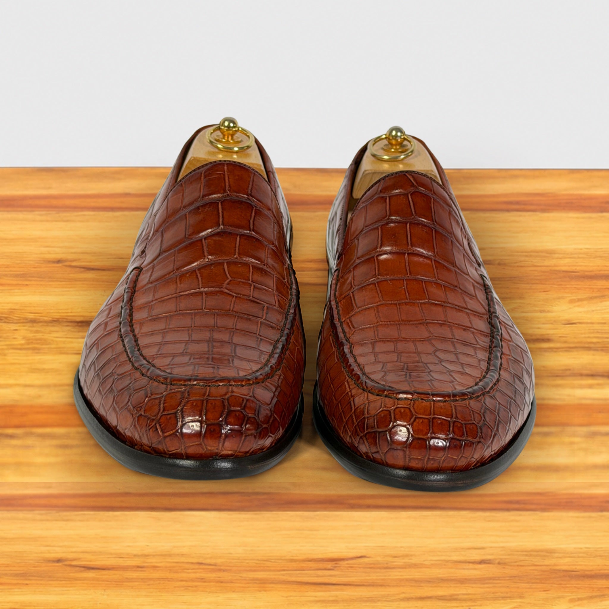 A pair of cognac crocodile-embossed leather loafers displayed on wooden shoe trees on a wooden surface against a light gray background.