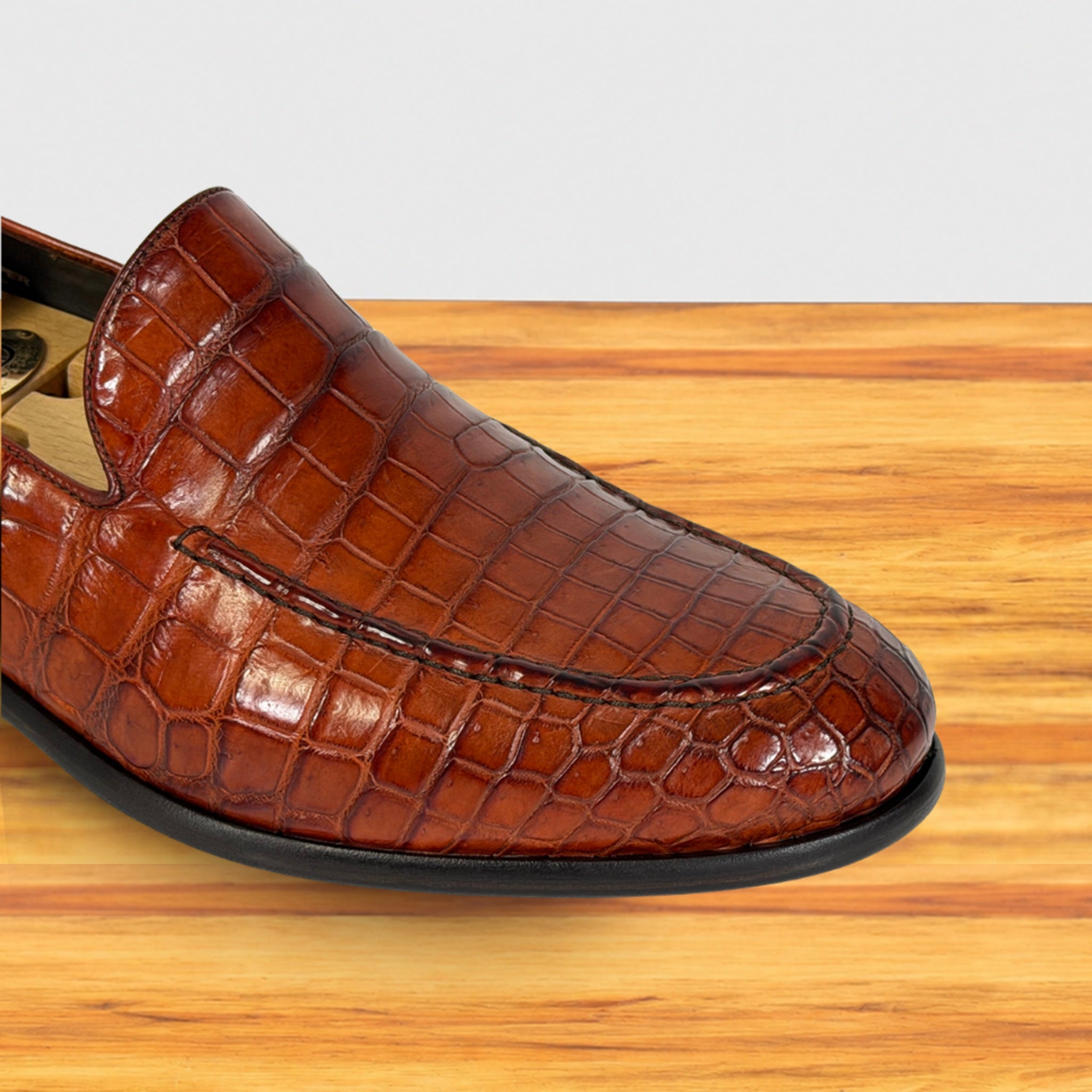A close-up of a cognac crocodile-embossed leather loafer shoe resting on a light wood surface against a light gray background.