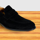 A black suede loafer shoe displayed on a wooden surface against a light gray background.