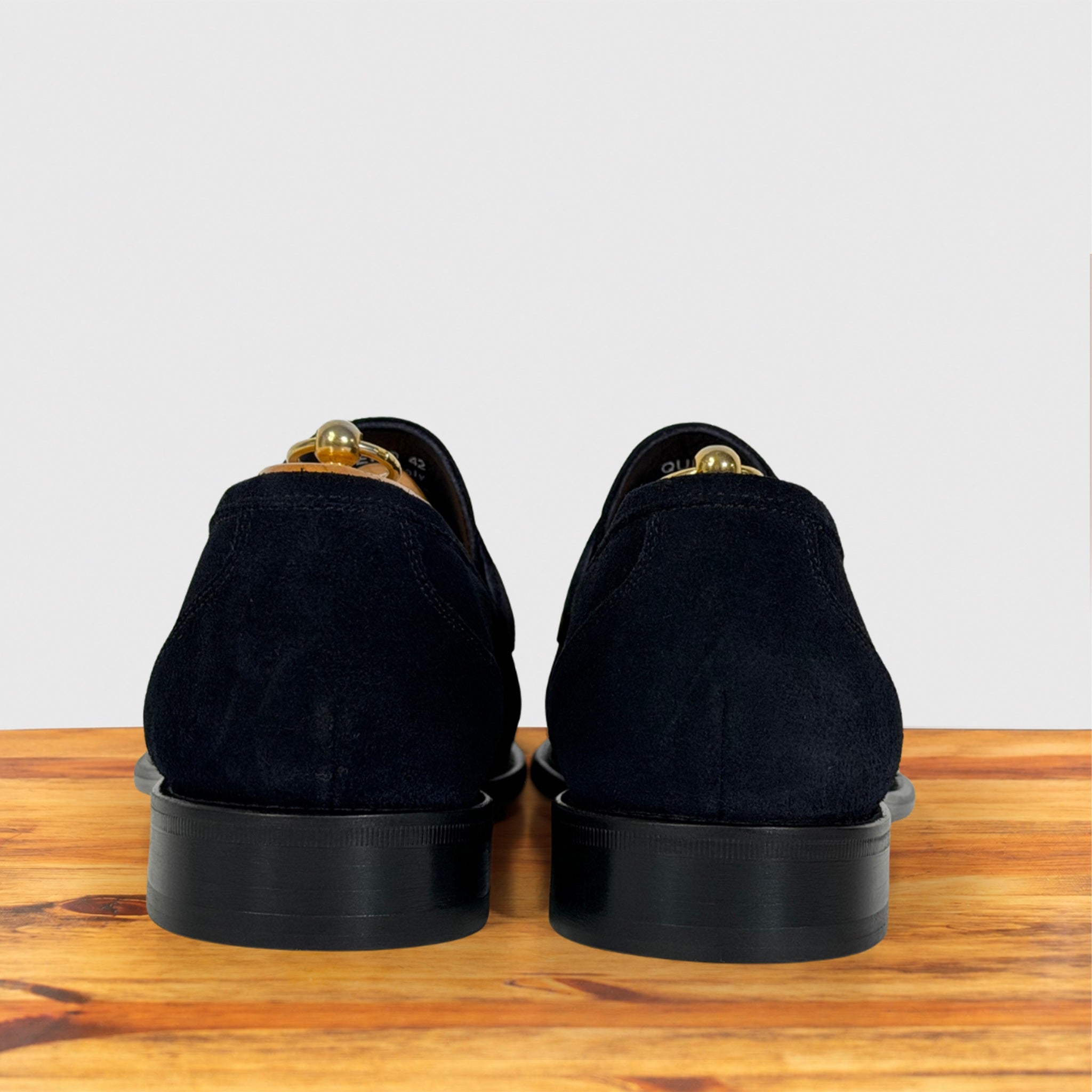 A pair of black suede loafers, shown from the back on a wooden surface against a light gray background.