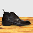 A dark brown leather lace-up ankle boot with a low stacked heel, shown in profile on a wooden surface against a light gray background.